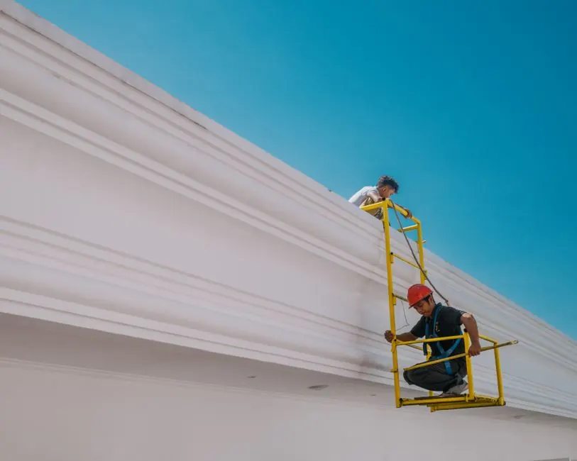 Two construction workers on a platform working on a white building under a clear sky.